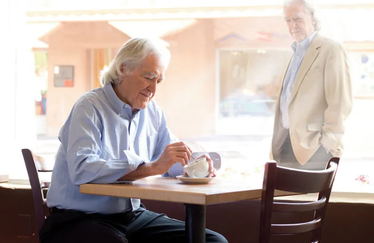 Dr. Larry Dossey gazes through a window at himself, seated in a cafe.