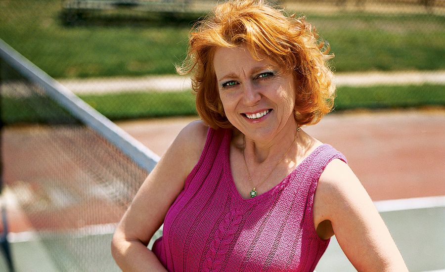 A smiling Noma Jewell leans on a tennis net while holding a racket.