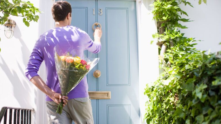 A man knocking on a door with a bouquet of flowers behind his back on a Spring day.