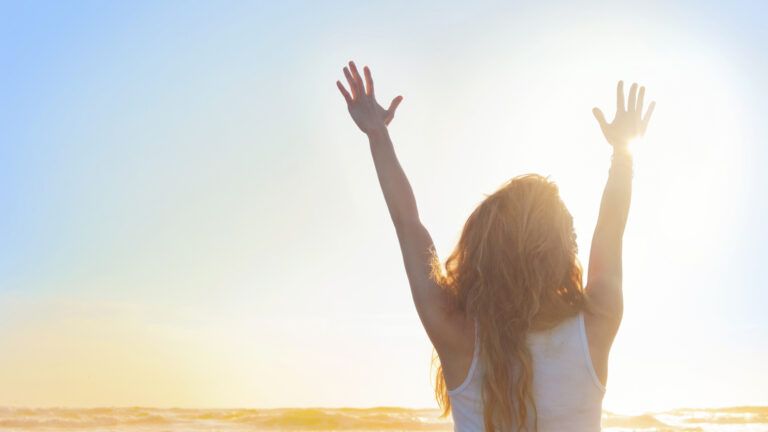 A woman with her hands raised in praise on a sun lit beach.