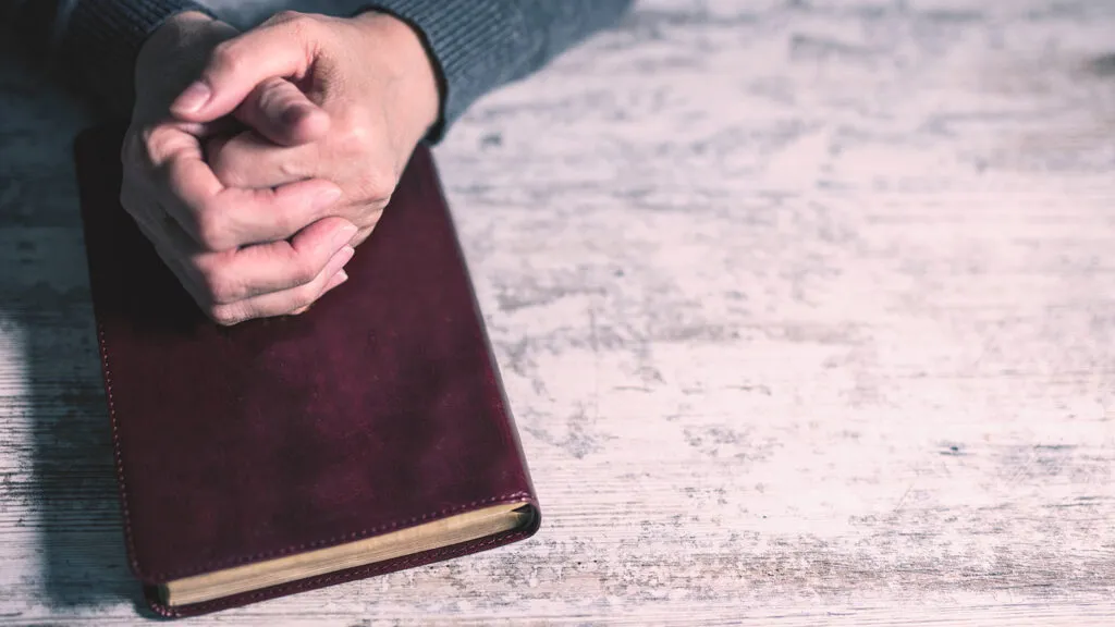 A man's hands clasped in prayer rest on a Bible