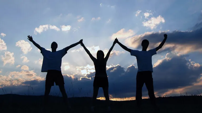 Three people stand facing the sunrise, arms upraised