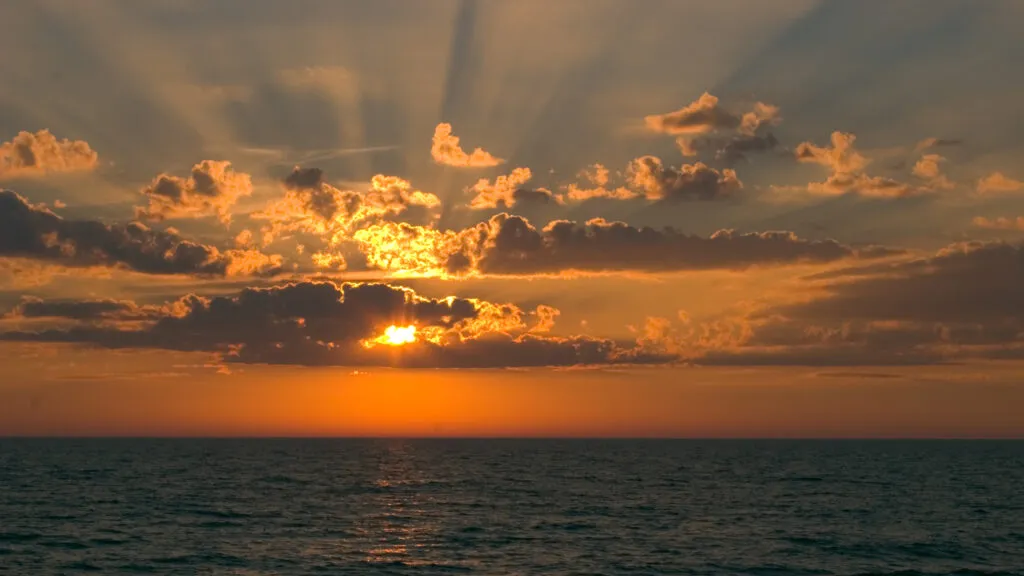 The Sun peeking from behind the clouds by a body of water.