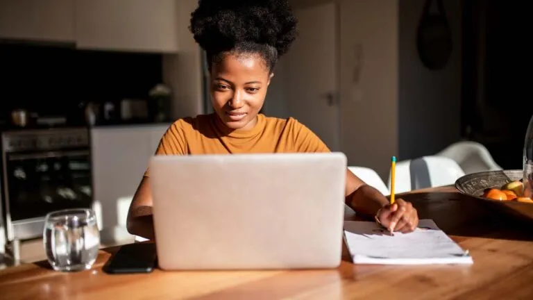 A woman using her laptop and writing down on paper; Getty Images