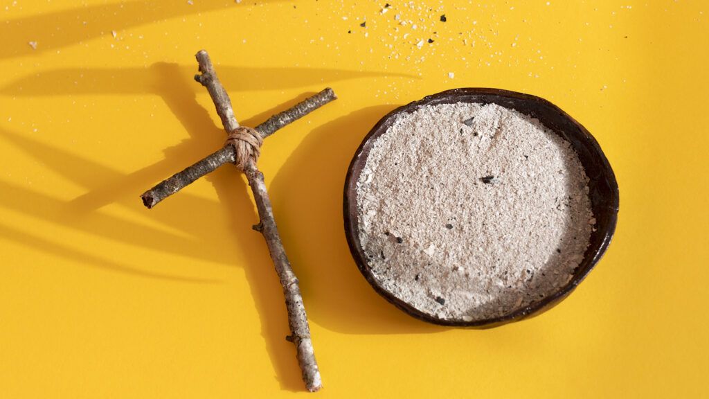 A wooden cross and ashes on a yellow background to show a Happy Ash Wednesday